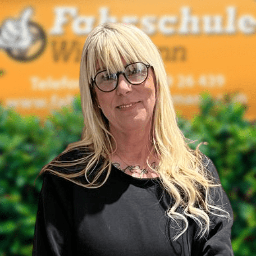 Blonde Frau mit Brille vor Fahrschule Wichmann Rastede Logo und grüner Hecke