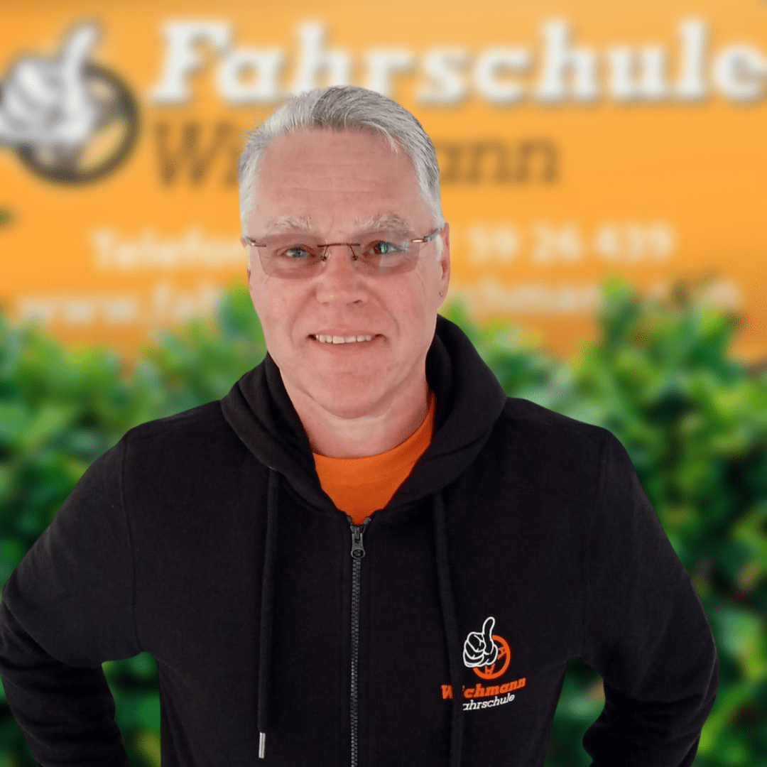 Fahrlehrer vor orangefarbener Wand mit Fahrschule Wichmann Logo in Rastede