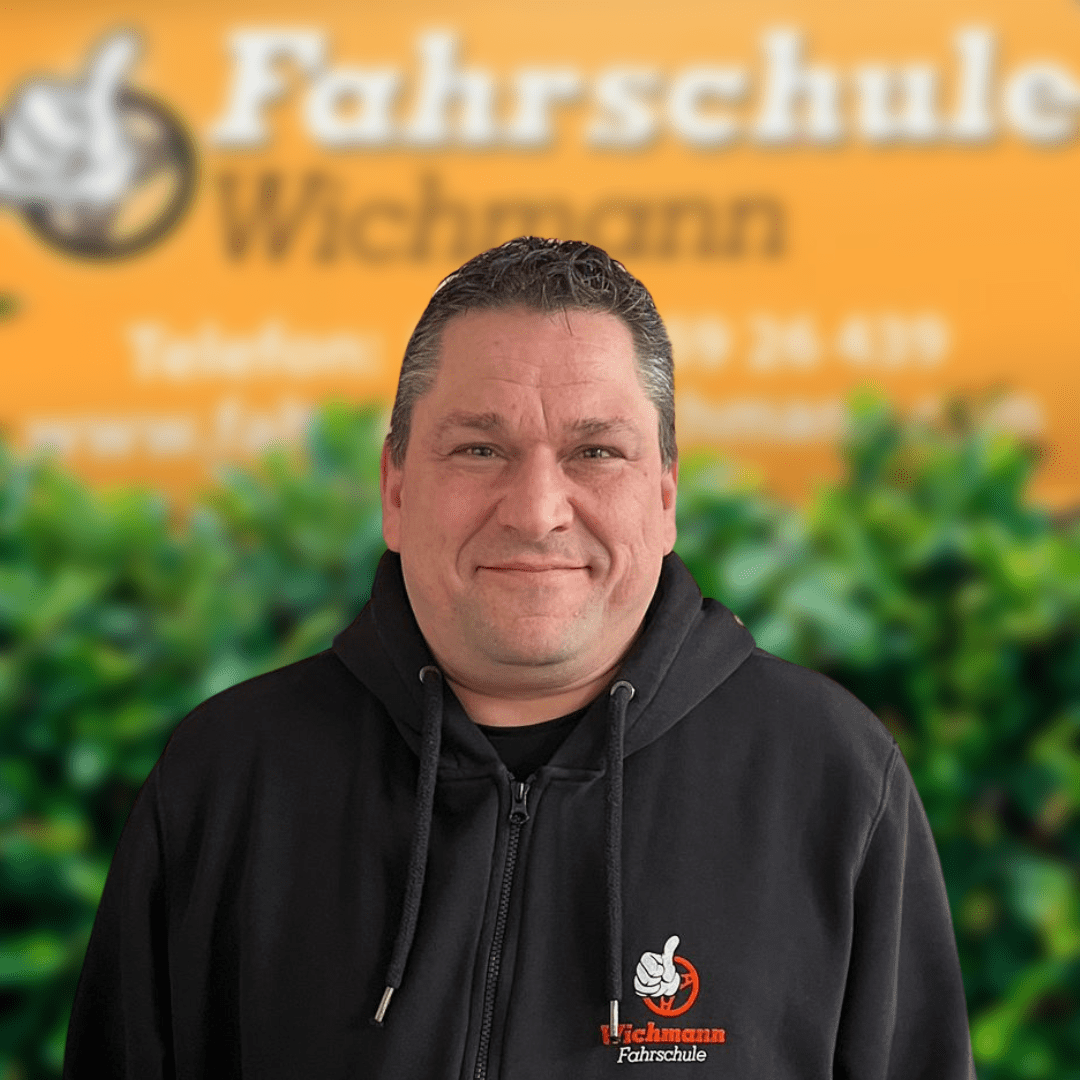 Fahrlehrer der Fahrschule Wichmann Rastede vor gelbem Schild mit Logo und Pflanzen