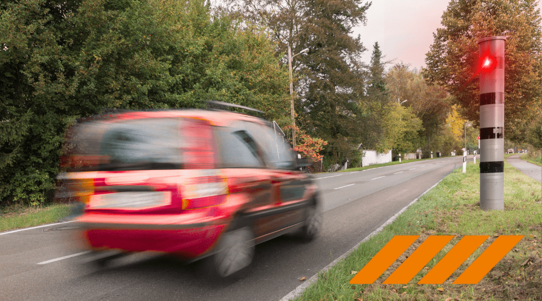 Blurred red car speeding along a tree-lined road, with a red traffic signal post nearby and orange warning stripes on the pavement.