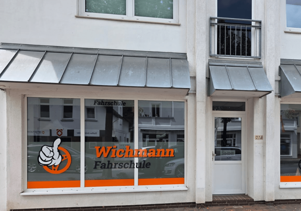 storefront of a driving school named 'Wichmann Fahrschule' with large windows, orange logo and text, and a thumbs-up graphic on the left window - A driving school shop front with glass windows displaying the orange 'Wichmann Fahrschule' sign and a large thumbs-up emblem - Exterior of a driving school building with tinted windows, orange accent stripe, and the word 'Fahrschule' visible in the window sign - Front of a building showing a driving school window display and a door to the right, featuring an orange logo and 'Wichmann Fahrschule' - Ground-floor storefront with metal awnings, signage reading 'Wichmann Fahrschule', and an illustrated thumbs-up logo on the left pane