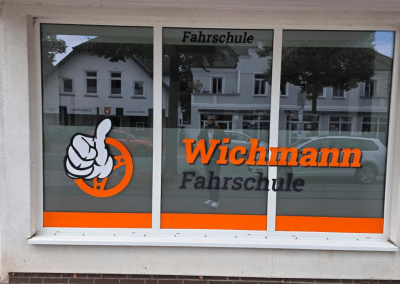 Fahrschule Wichmann Fenster mit orangefarbenem Logo und Daumenhoch in Rastede